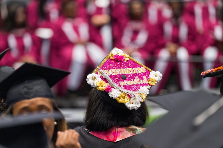 A student wearing a commencement hat decked with flowers, which says 'Nonetheless She Persisted.'