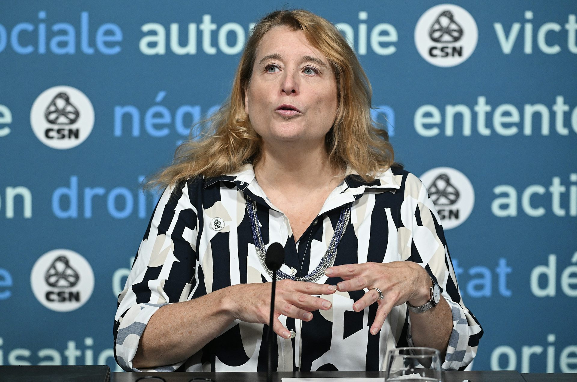 A middle-aged white woman in a black and white blouse speaks into a microphone from behind a table