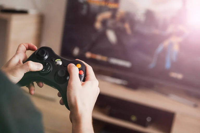 A pair of hands using a video game controller to play a fighting game on a television in the background