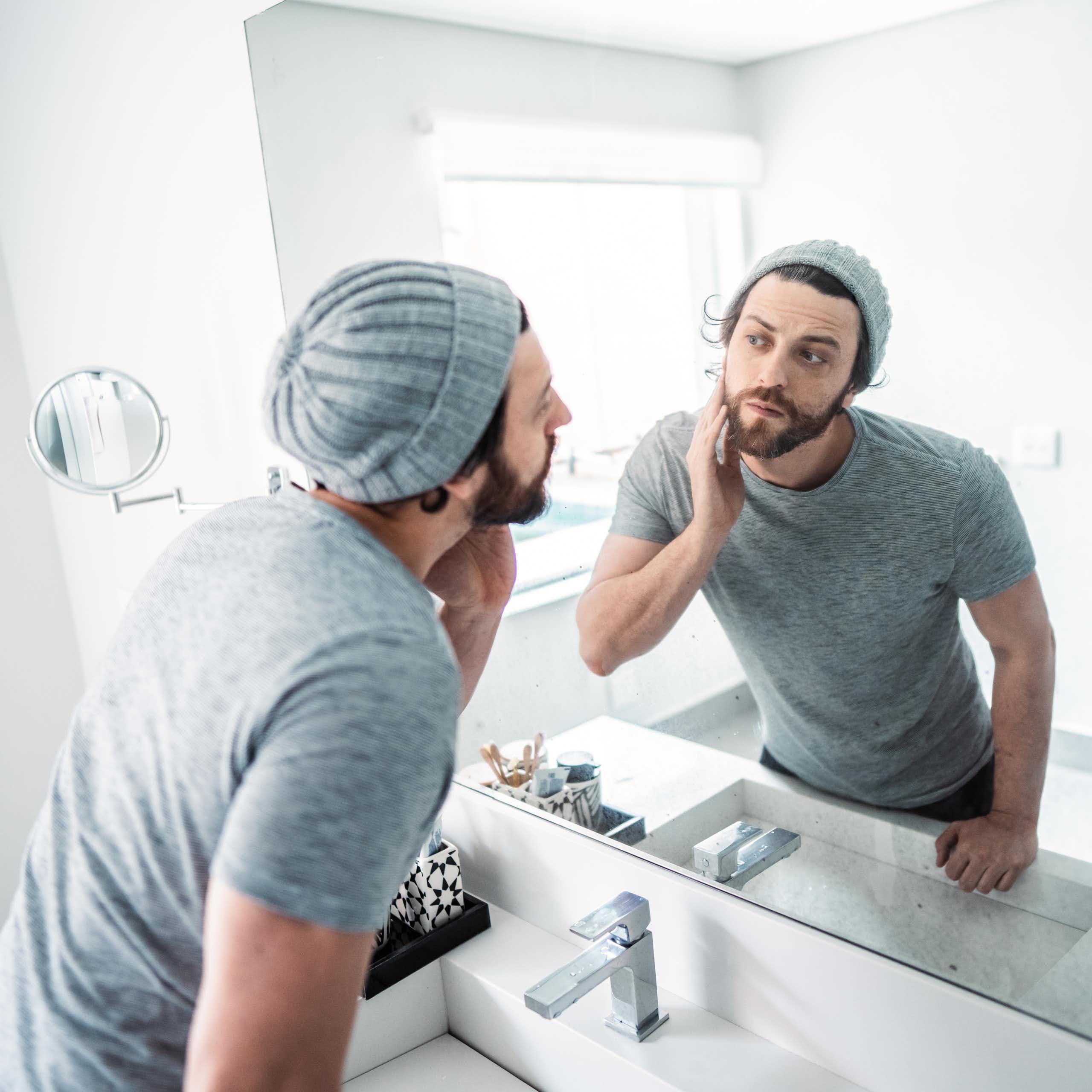 a young bearded man wearing a knit cap and t-shirt touches his face as he looks at himself in a bathroom mirror