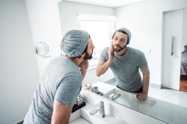a young bearded man wearing a knit cap and t-shirt touches his face as he looks at himself in a bathroom mirror