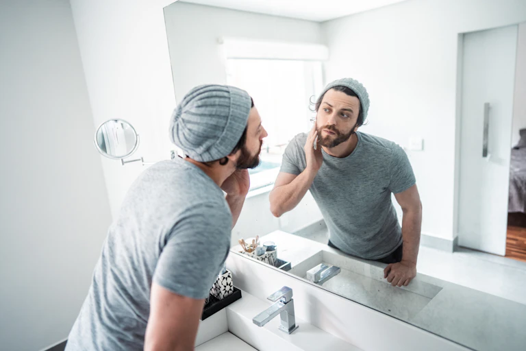 a young bearded man wearing a knit cap and t-shirt touches his face as he looks at himself in a bathroom mirror