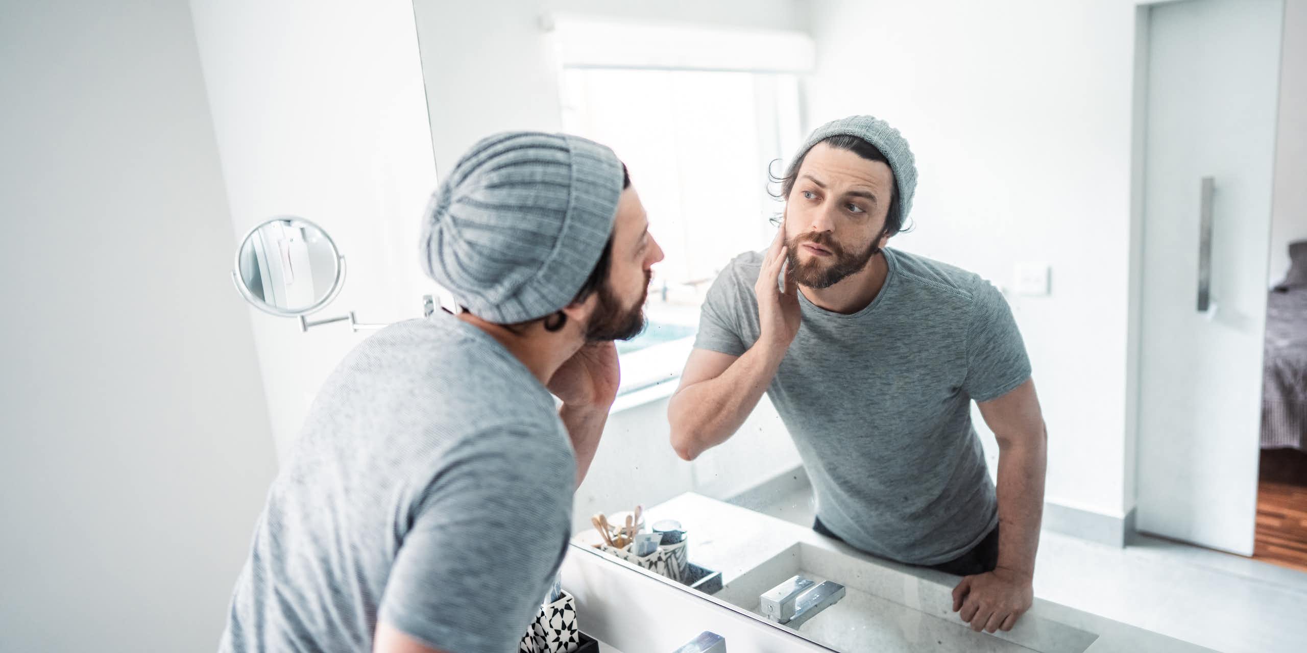 a young bearded man wearing a knit cap and t-shirt touches his face as he looks at himself in a bathroom mirror