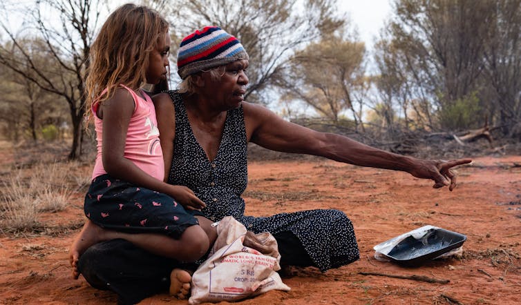 An older Aboriginal woman sits on the ground in the outback, directing a young Aboriginal girl, who sits on her lap