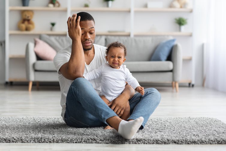 man seated on floor with his hand to his head, supporting a crying baby