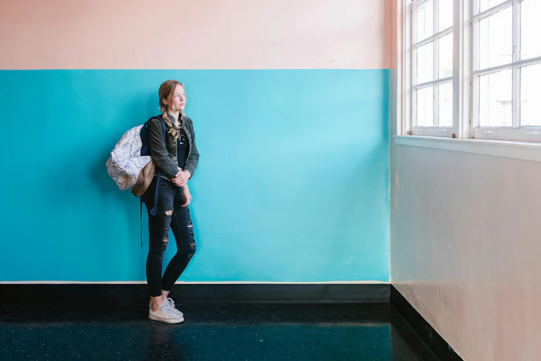 A young woman in jeans and a backpack leans against a blue and pink painted wall. She looks out of a large window.