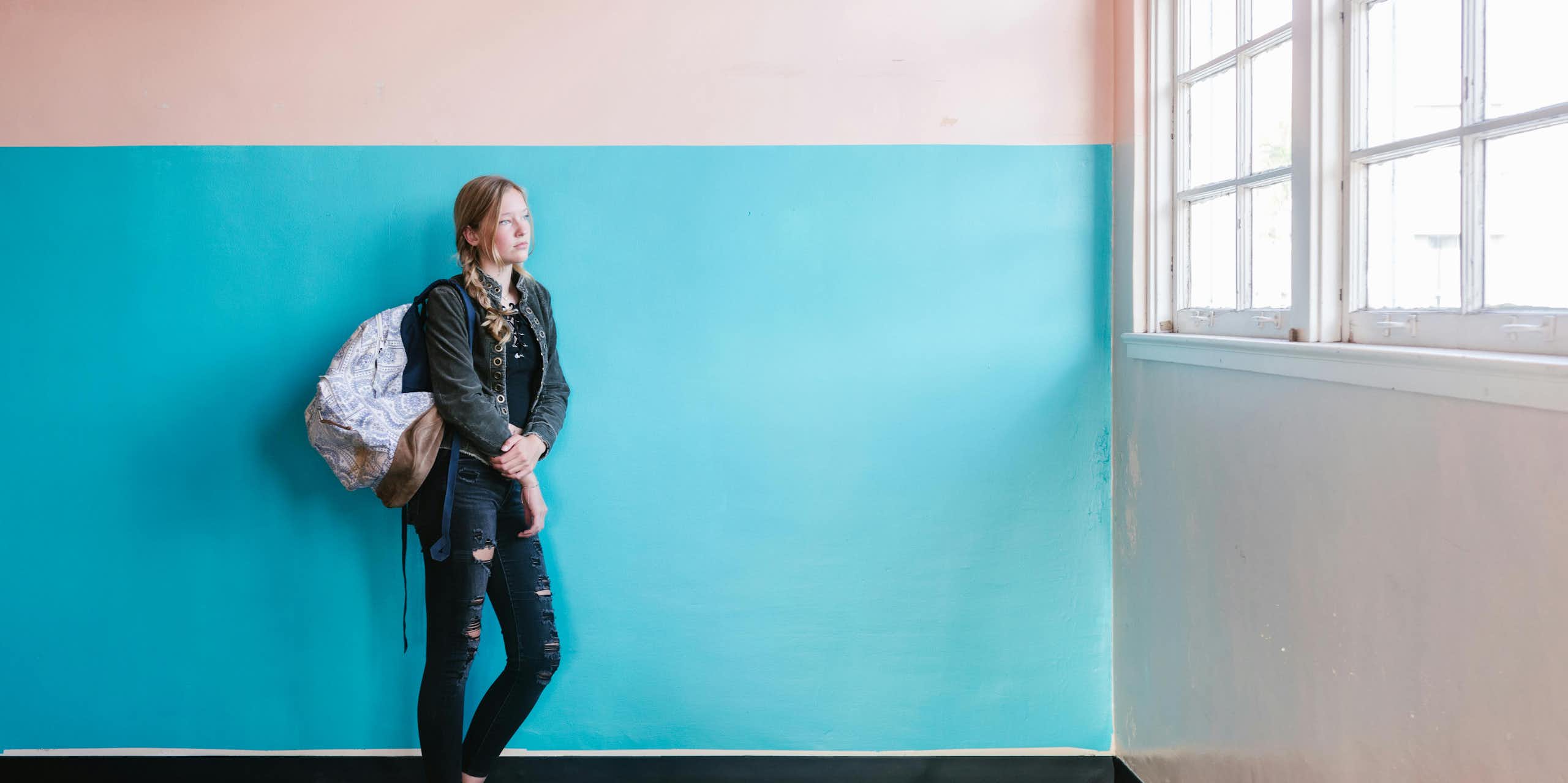 A young woman in jeans and a backpack leans against a blue and pink painted wall. She looks out of a large window. 