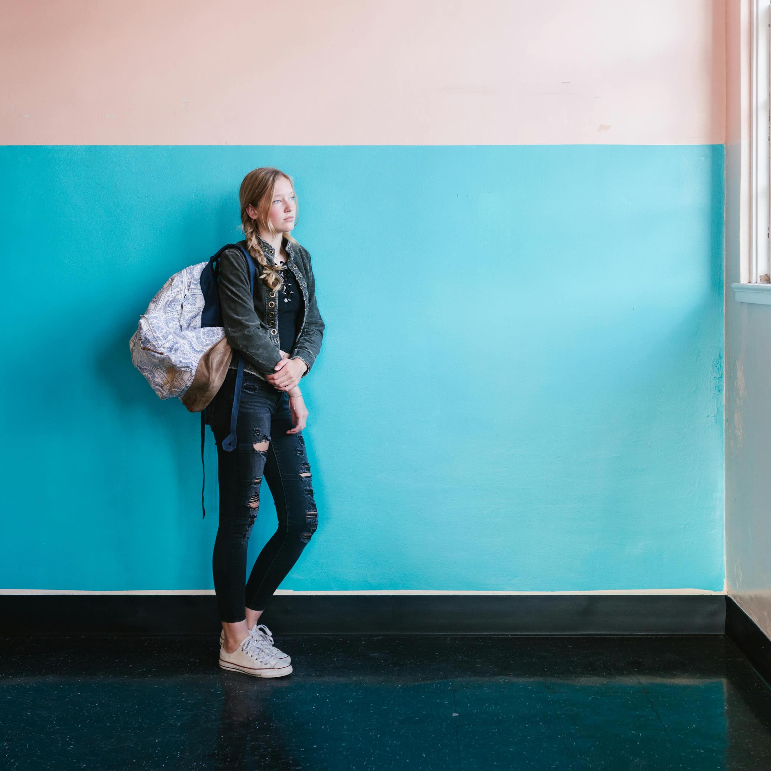 A young woman in jeans and a backpack leans against a blue and pink painted wall. She looks out of a large window. 