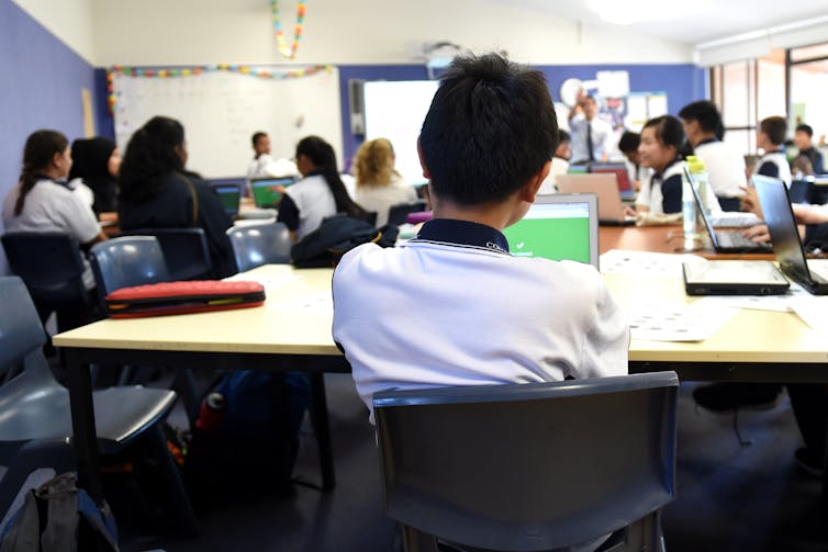 Students sit at desks in a classroom, a teacher stands at the front.