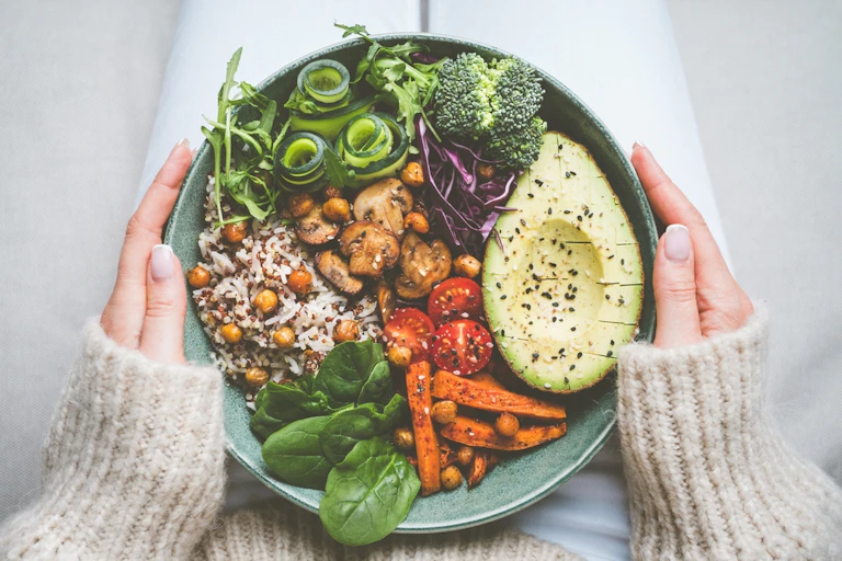 Woman holding bowl of vegan food (avocado, veggies, rice) on lap