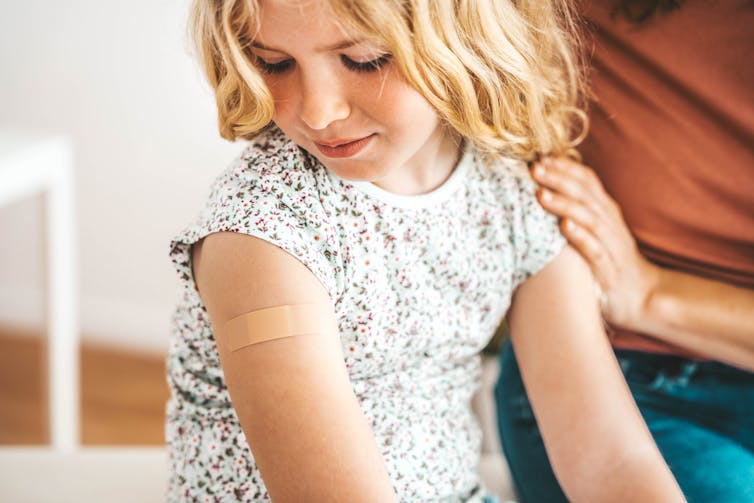 A girl looking down at a bandaid on her upper arm.