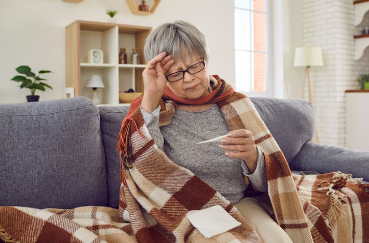 A senior woman sitting on the couch wrapped in a blanket and looking at a thermometer.