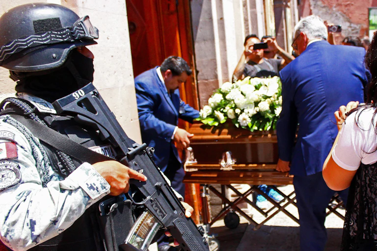 An armed guard standing outside a cathedral as a coffin is carried through the door.