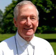 A grey haired white man, Raymond Briggs smiling in a park on a summer's day.