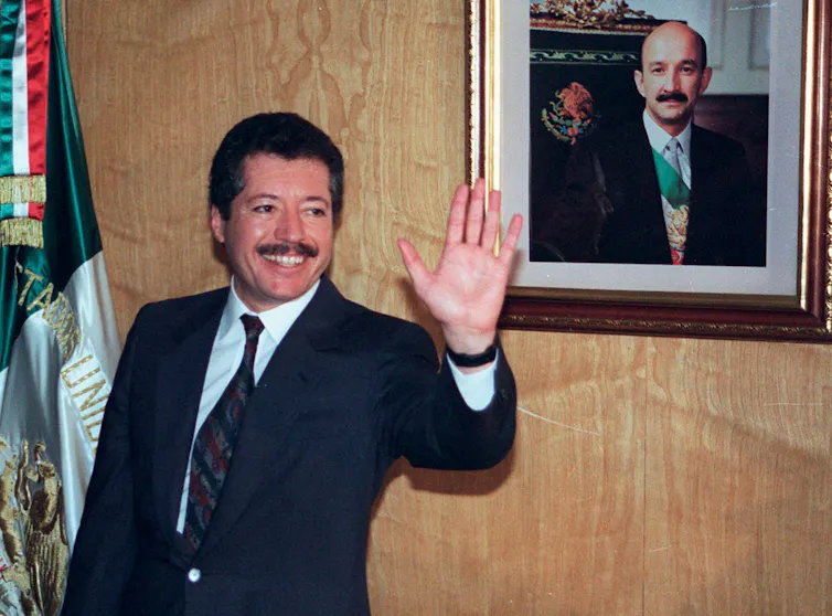 A man with a moustache, flanked by a Mexican national flag and a framed portrait of suited man, waving to reporters.