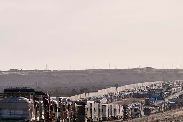 Two very long lines of trucks congest a highway, with a white wall and grey sky in the background.