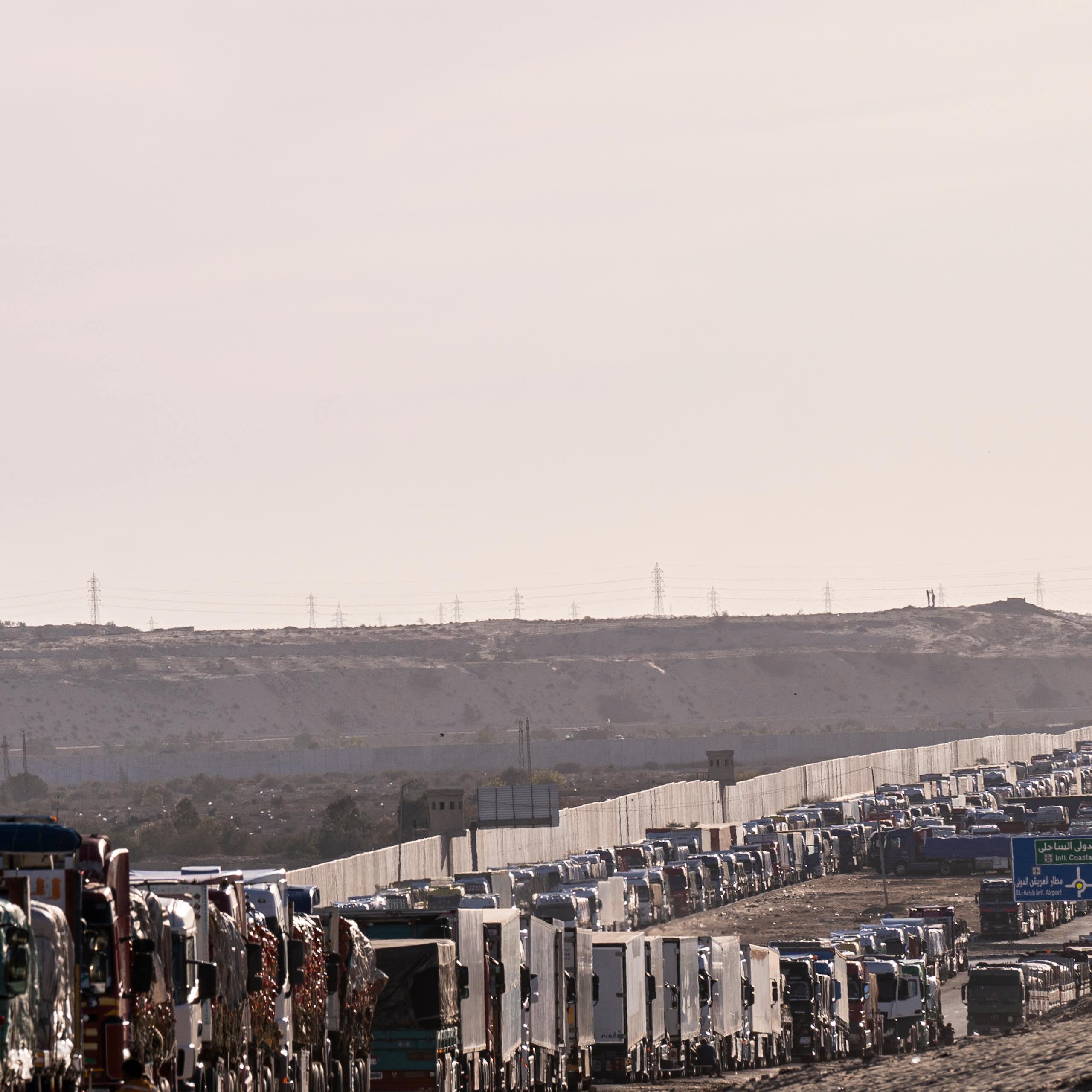 Two very long lines of trucks congest a highway, with a white wall and grey sky in the background.