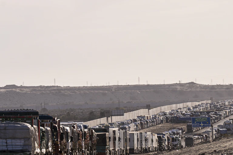 Two very long lines of trucks congest a highway, with a white wall and grey sky in the background.