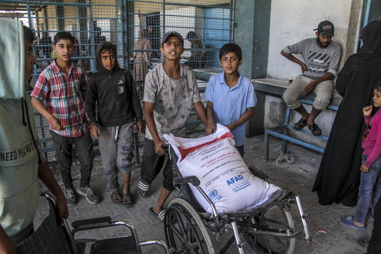 Four boys, who appear to be teenagers, look at a camera and push a wheelchair that has a large white bag on it.