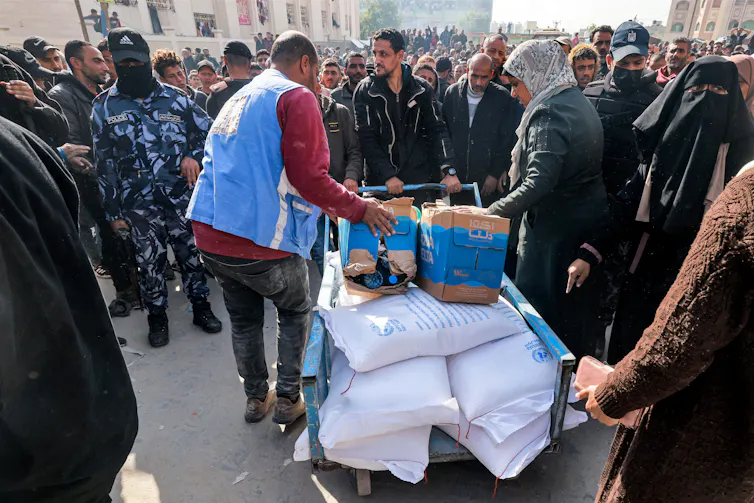 A man in a blue vest stands over a cart that has large white bags and cardboard boxes on it. Many men, including one in a blue camp uniform, surround him.
