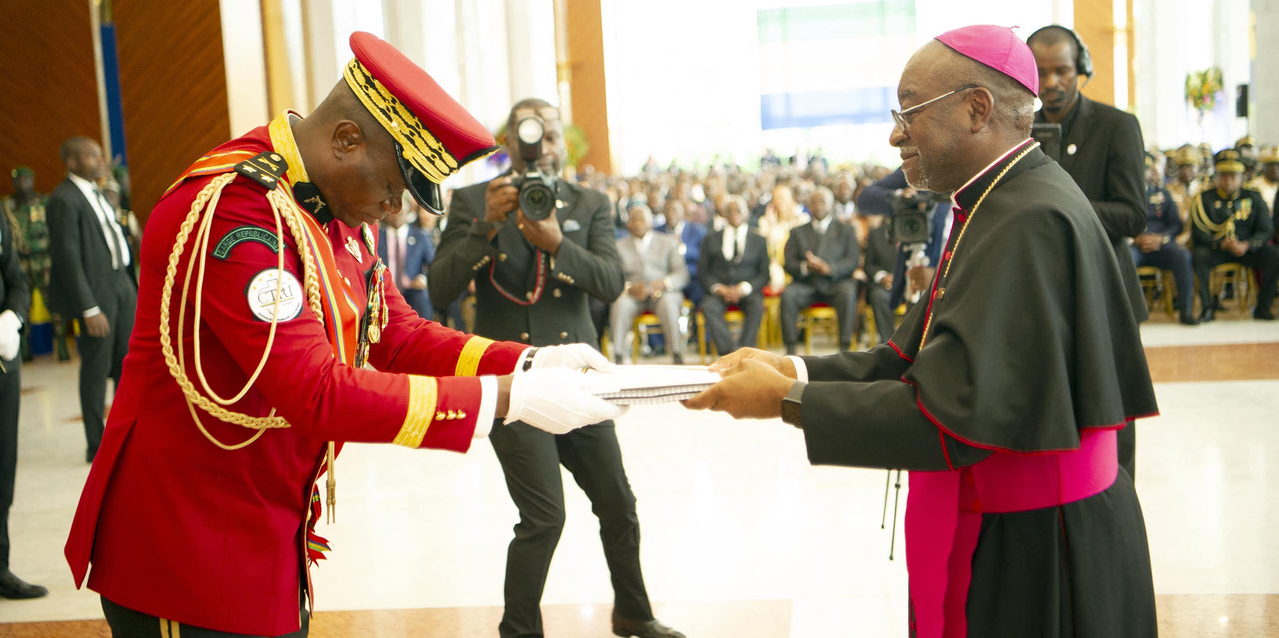 A man wearing ceremonial military uniform takes a bow while receiving a document from another man wearing a cassock.