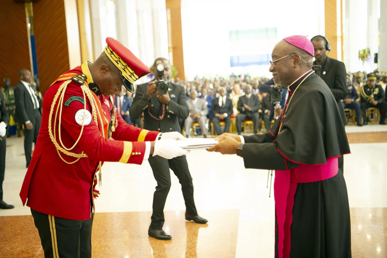 A man wearing ceremonial military uniform takes a bow while receiving a document from another man wearing a cassock.