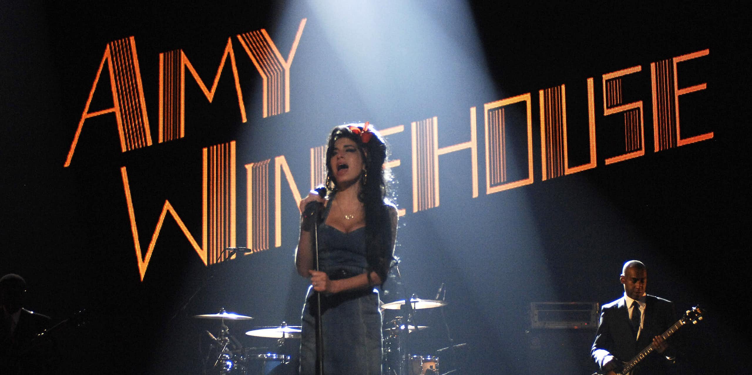 Late singer Amy Winehouse, whose name is displayed in lights, performs on a stage with musical instruments and a guitar player behind her.
