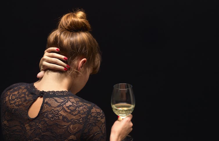 Adult woman looking away from the camera, holding a glass of white wine in one hand and pressing her left hand against her neck.