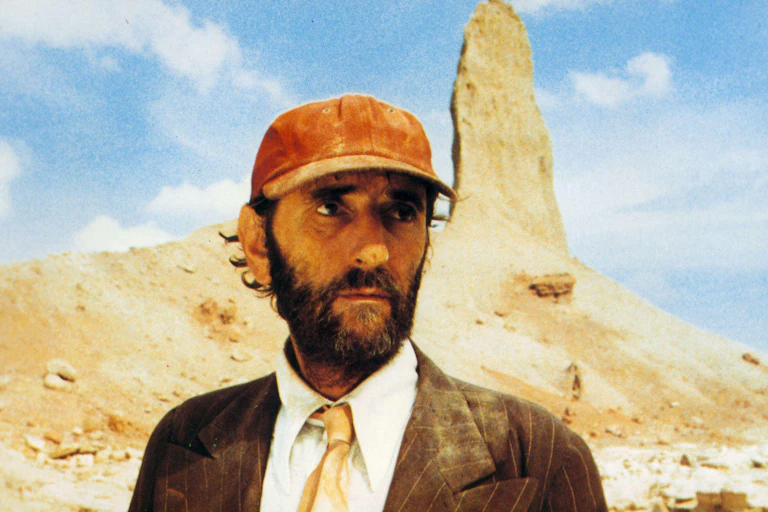 A man in a dusty suit and red baseball cap wandering the deserts of Texas.