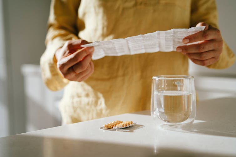 Person holding a patient information leaflet over a blister pack of pills and a glass of water on a counter
