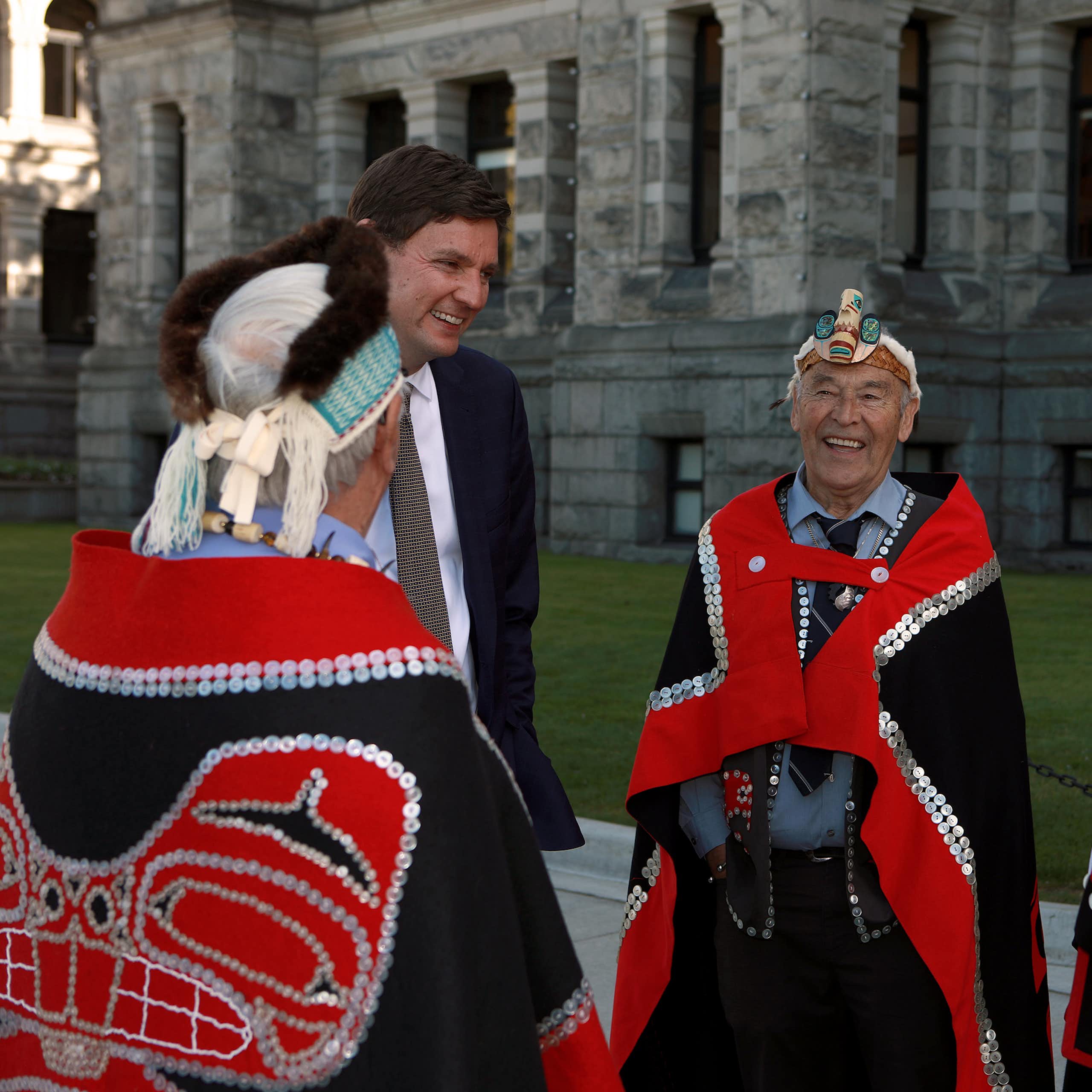 A man in a suit smile and he talks with Indigenous men in red and blue ceremonial garb who are also smiling.