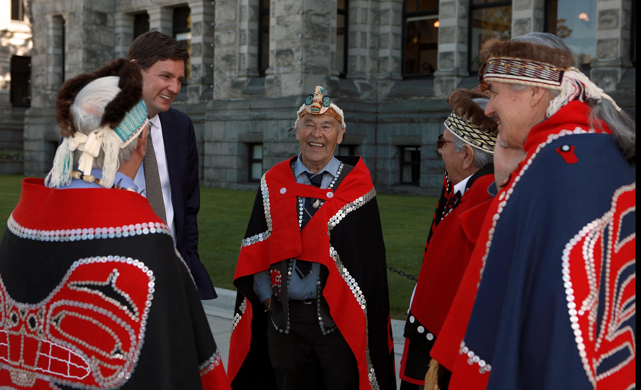 A man in a suit smile and he talks with Indigenous men in red and blue ceremonial garb who are also smiling.