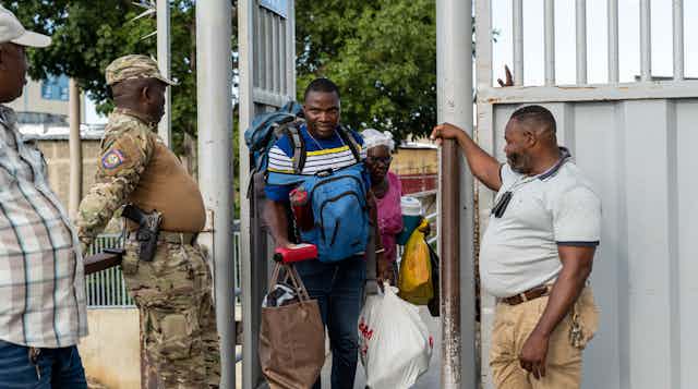 A man laden with bags walks through a metal door flanked by two security guards.