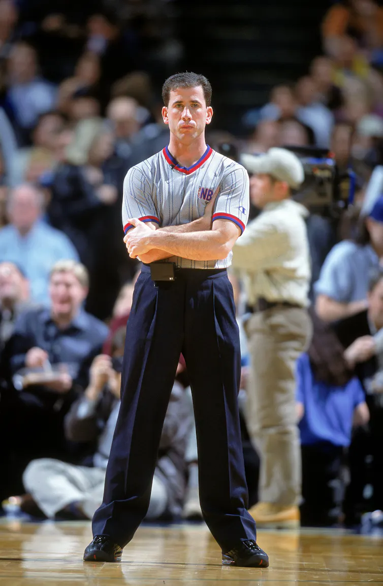 A man stands on a basketball court with a wide stance and arms crossed. Spectators can be seen behind him.