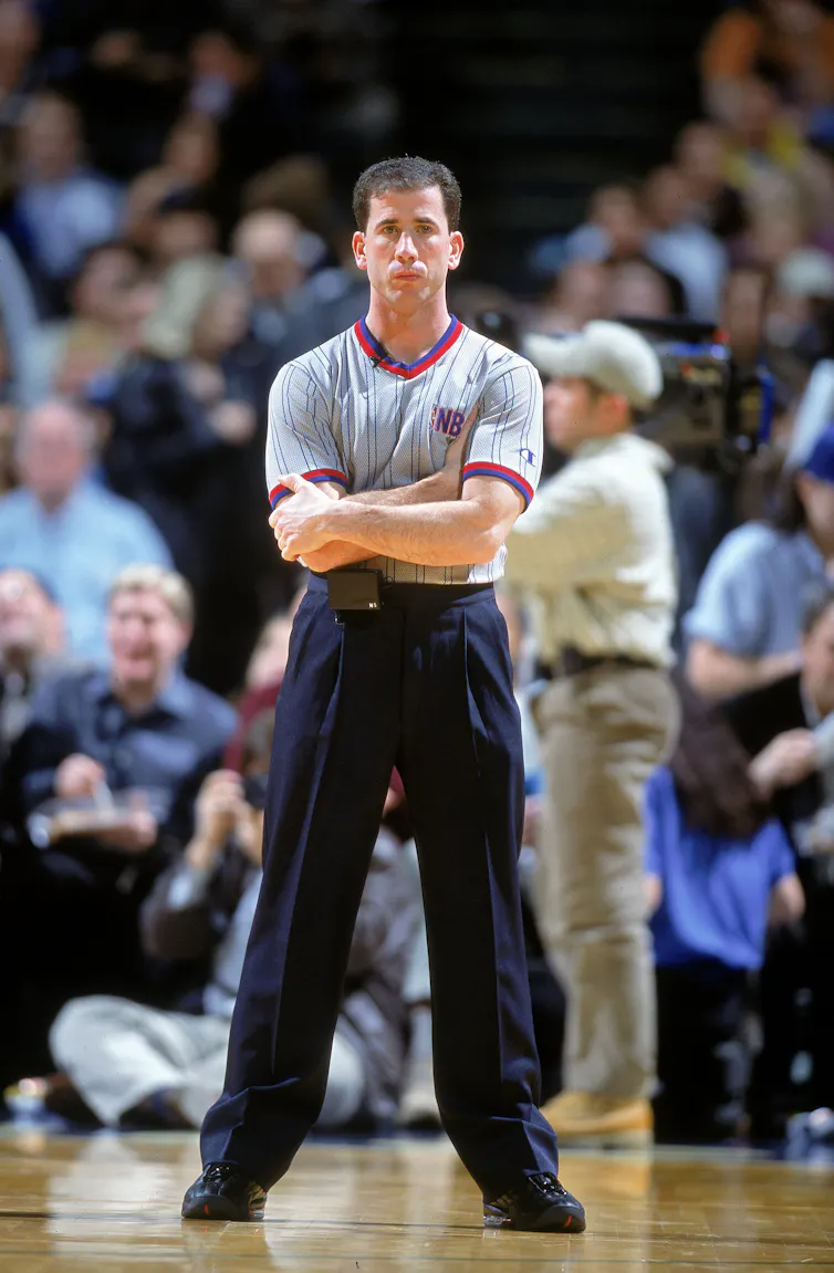 A man stands with a wide stance and crossed arms on a basketball court. Spectators are visible behind him.
