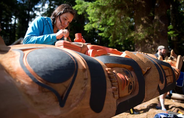 A woman with a paint brush works on a large totem pole.