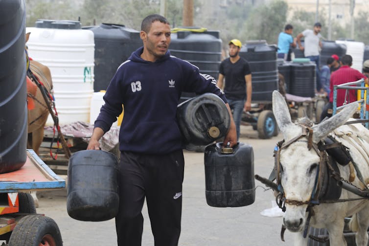 A man carrying three large plastic jugs walks next to a donkey cart