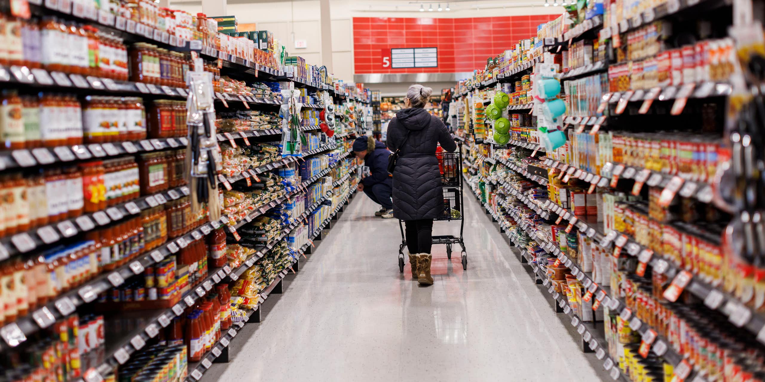 A person seen from behind pushing a grocery cart down an aisle in a supermarket