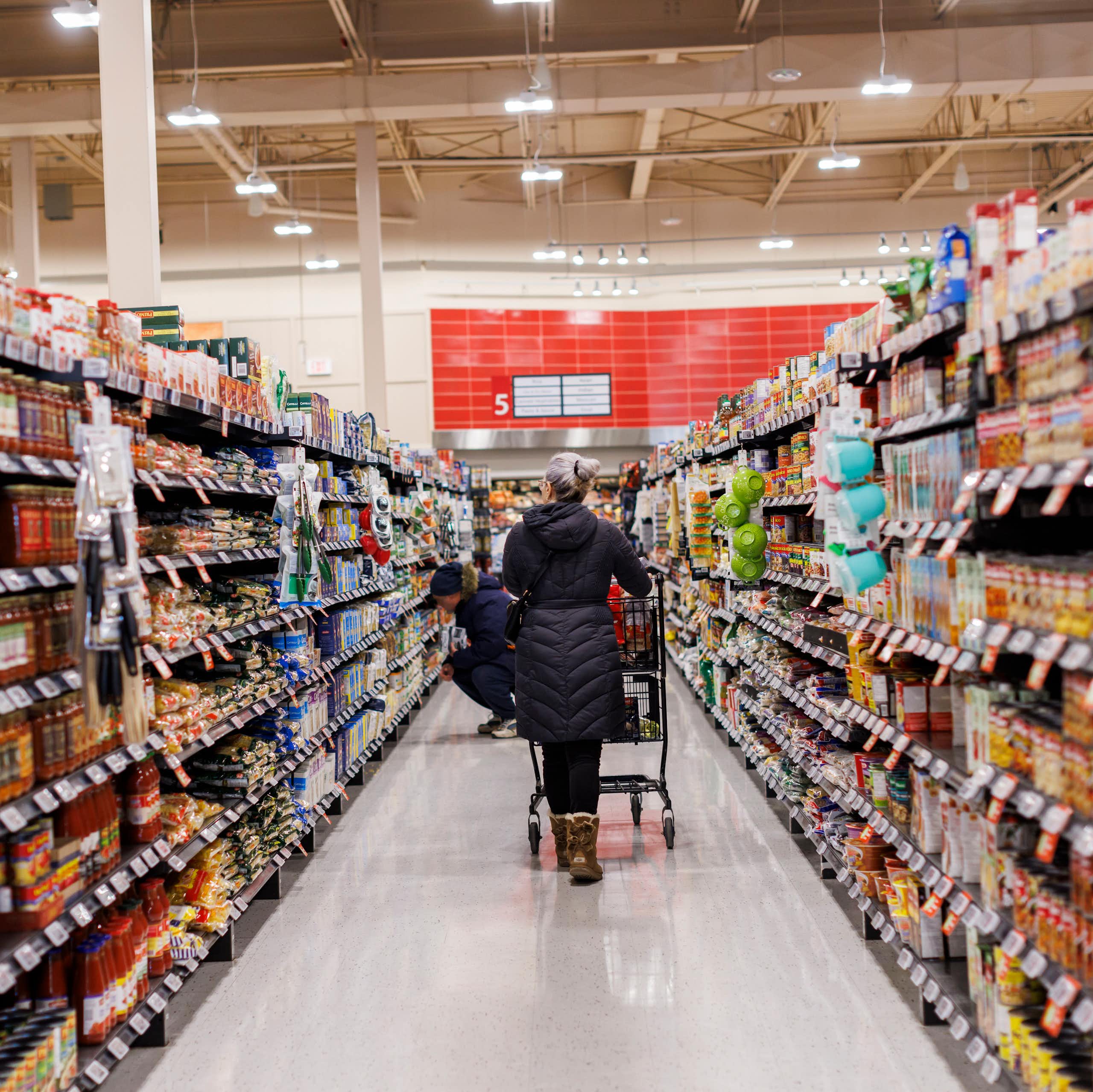 A person seen from behind pushing a grocery cart down an aisle in a supermarket