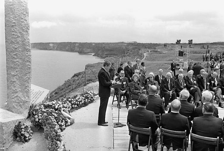 Backed by a monument and the sea, a man speaks to a crowd.