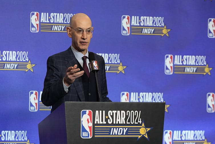 A middle-aged bald man in glasses speaks from behind a podium emblazoned with the NBA logo and signage that says All-Star 2024