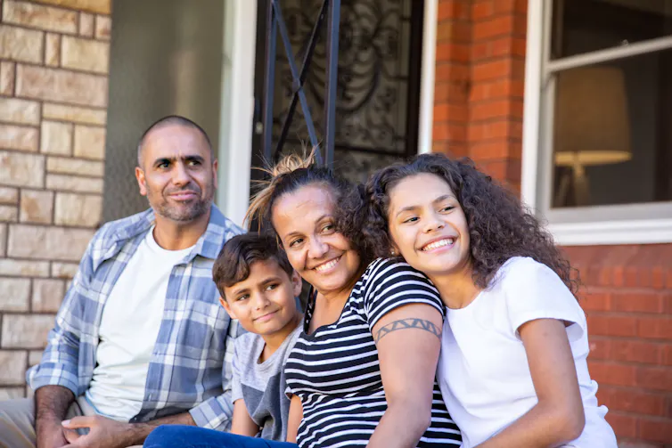An Indigenous family of father, young son, mother and older daughter sit on a doorstep happily