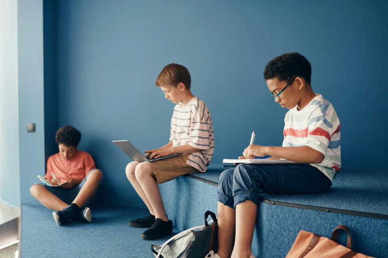 Three boys sit in a row, writing. Two do so with pens and notebooks. One types on a laptop.