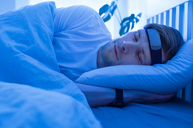 A man sleeping in a blue lit room with a smart headband and a wristwatch.