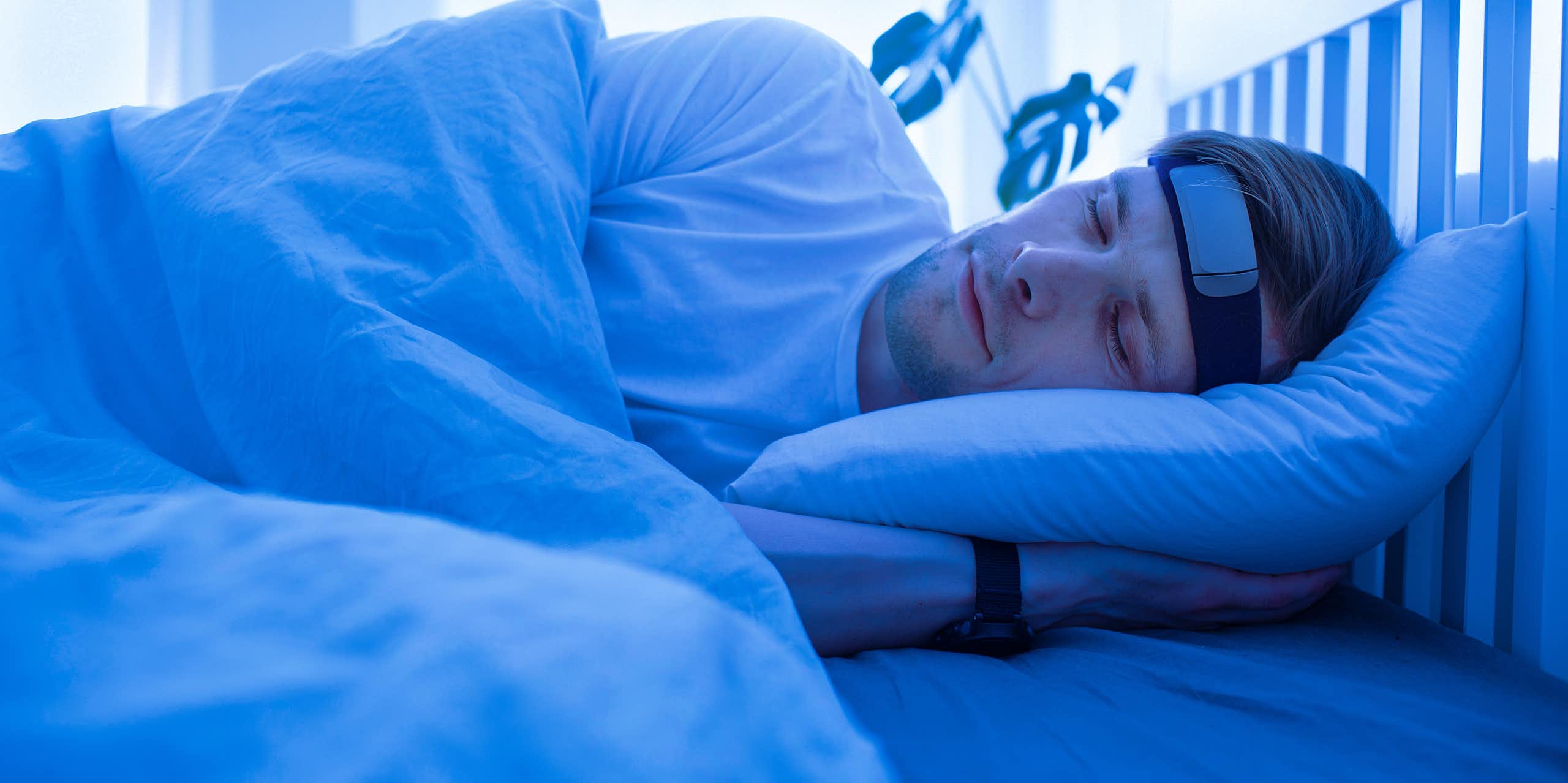 A man sleeping in a blue lit room with a smart headband and a wristwatch.