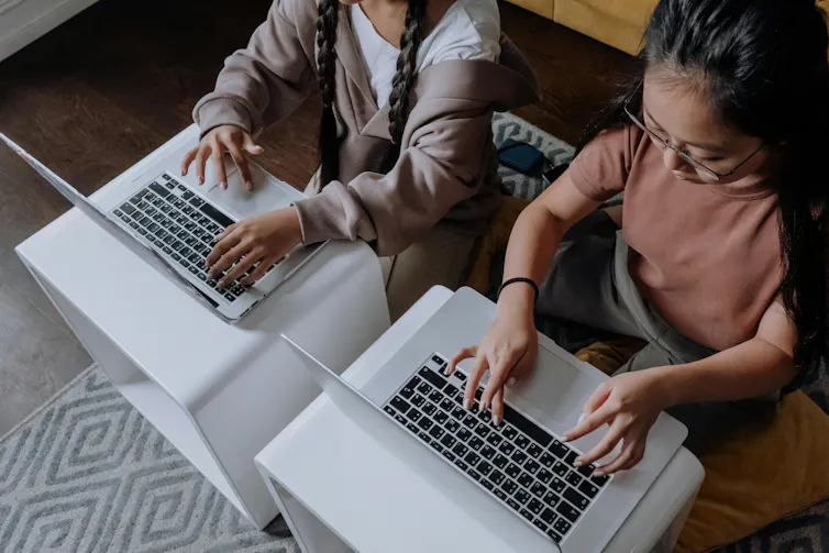 Two children sit side by side, using laptops on small tables.
