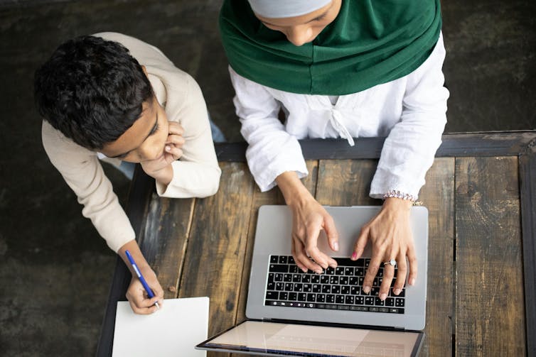 A woman types on a laptop. A boy sits next to her, watching. He has a pen and paper.