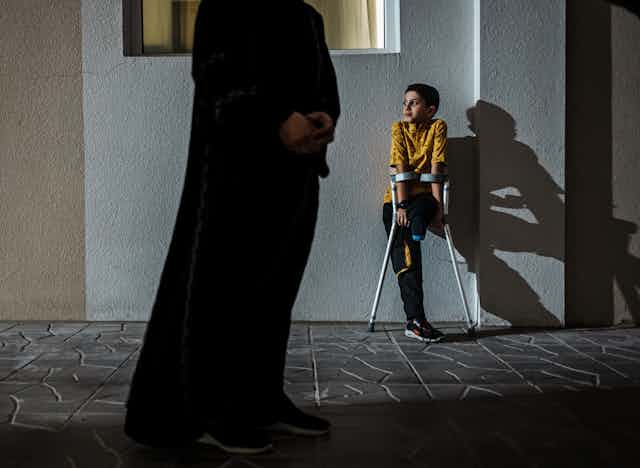 Un joven sin una pierna utiliza muletas mientras se apoya en una pared. En primer plano, un adulto sin rostro.