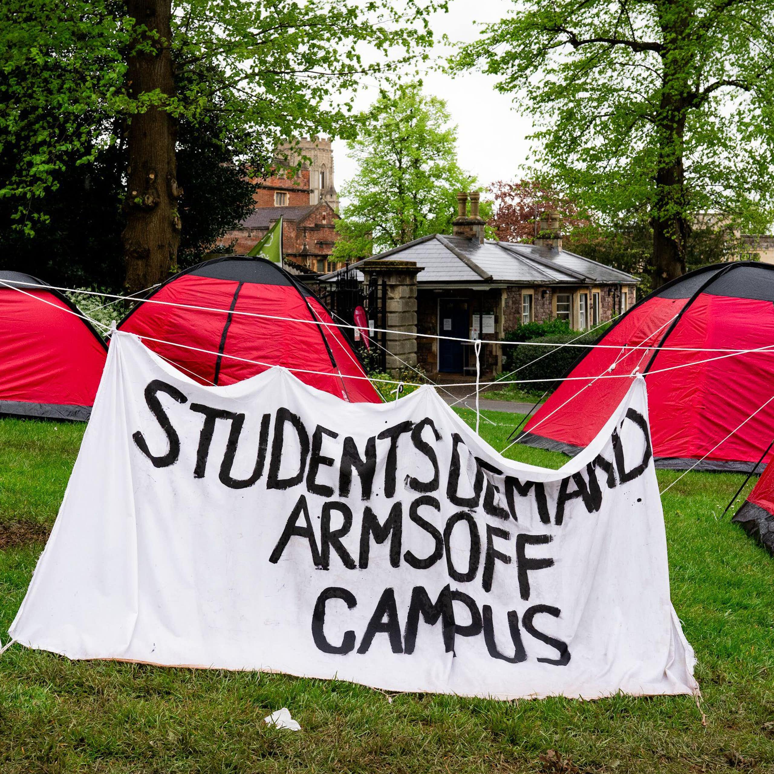 Red camping tents on a green lawn, with a large hand painted sign reading 'students demand arms off campus'