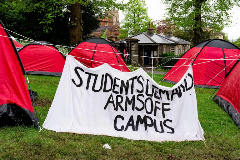 Red camping tents on a green lawn, with a large hand painted sign reading 'students demand arms off campus'